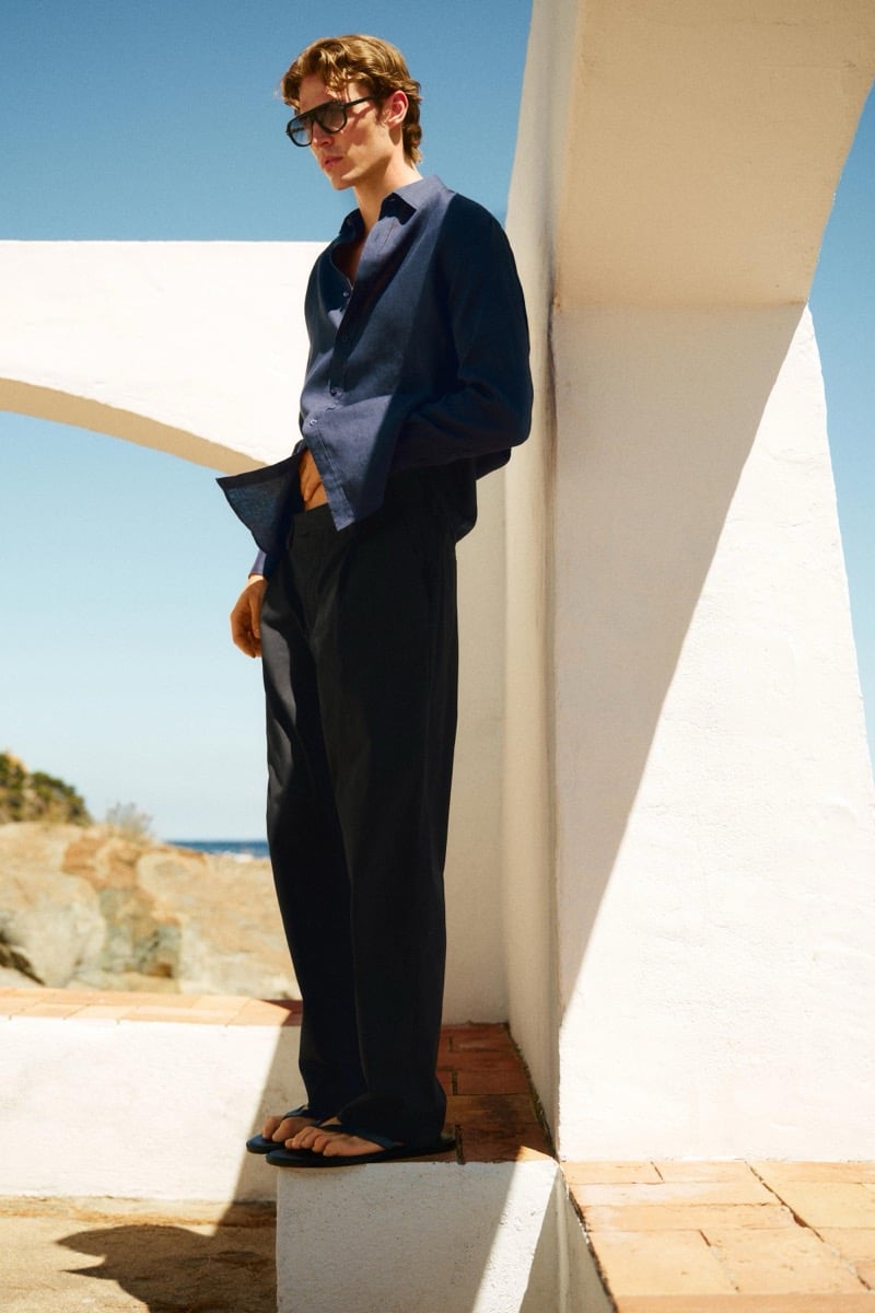 Callum Simpson stands on a stone ledge above the coast in an open-collar navy shirt, tailored trousers, and flip-flop sandals, sunglasses shading his eyes