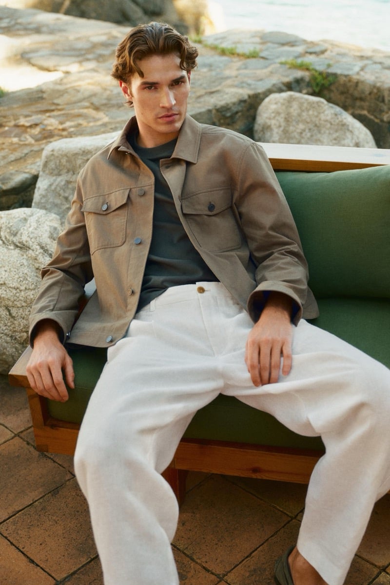 Callum Simpson sits on an outdoor sofa in a taupe field jacket over a teal T-shirt and ivory linen trousers, rocky coastline behind him