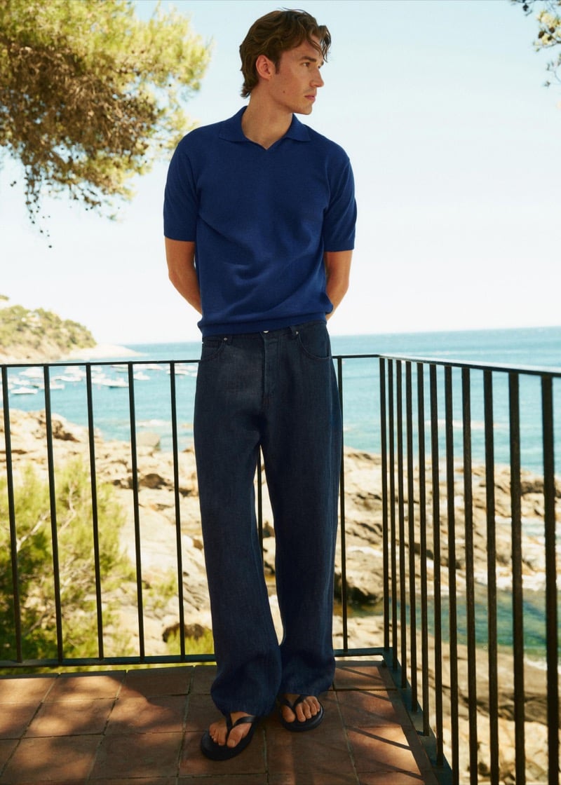 Callum Simpson stands on a balcony overlooking a marina in a cobalt knit polo and dark denim trousers, flip-flop sandals on his feet