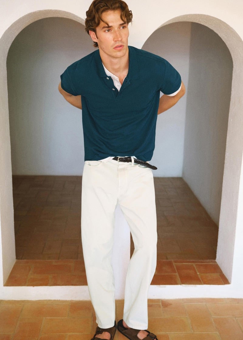 Callum Simpson stands in a whitewashed archway in a teal pique polo with white trim, ivory trousers, and a dark woven belt
