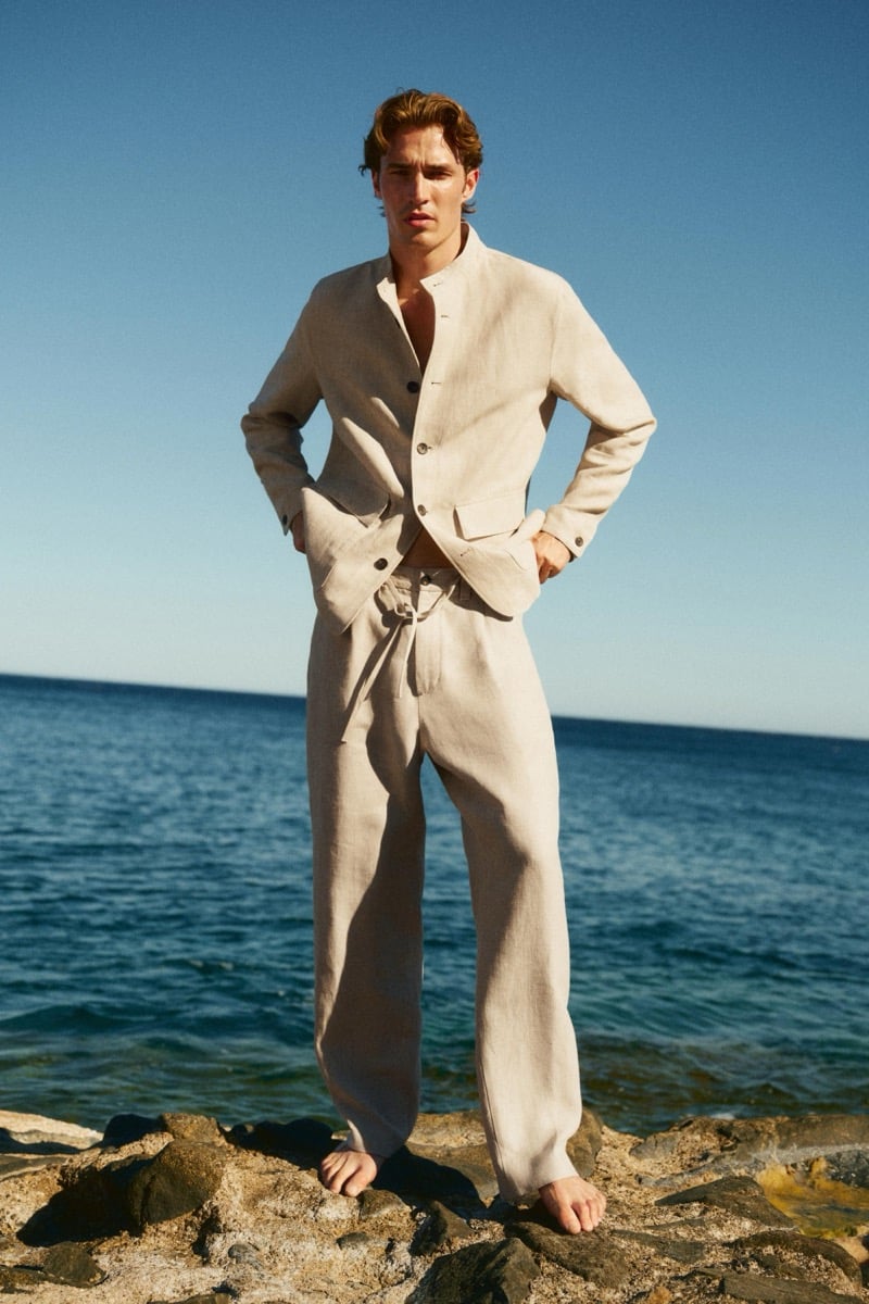 Callum Simpson stands barefoot on coastal rocks in an ivory linen suit with a mandarin collar, hands in his pockets