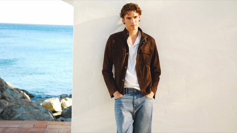 Callum Simpson leans against a whitewashed wall by the sea in a chocolate suede jacket over a white polo and light-wash jeans