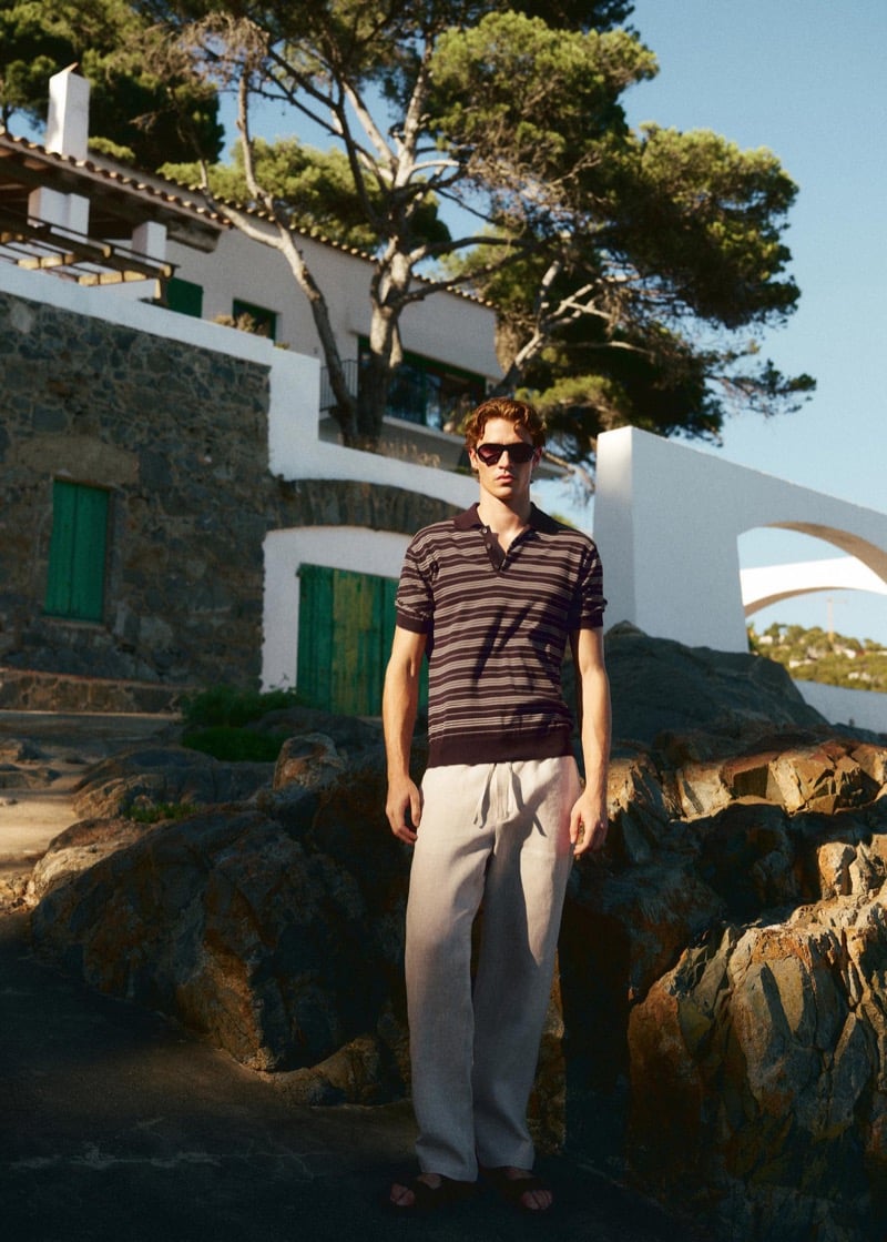 Callum Simpson stands before a whitewashed Mediterranean house in a burgundy and taupe striped polo with stone-colored linen trousers and sandals