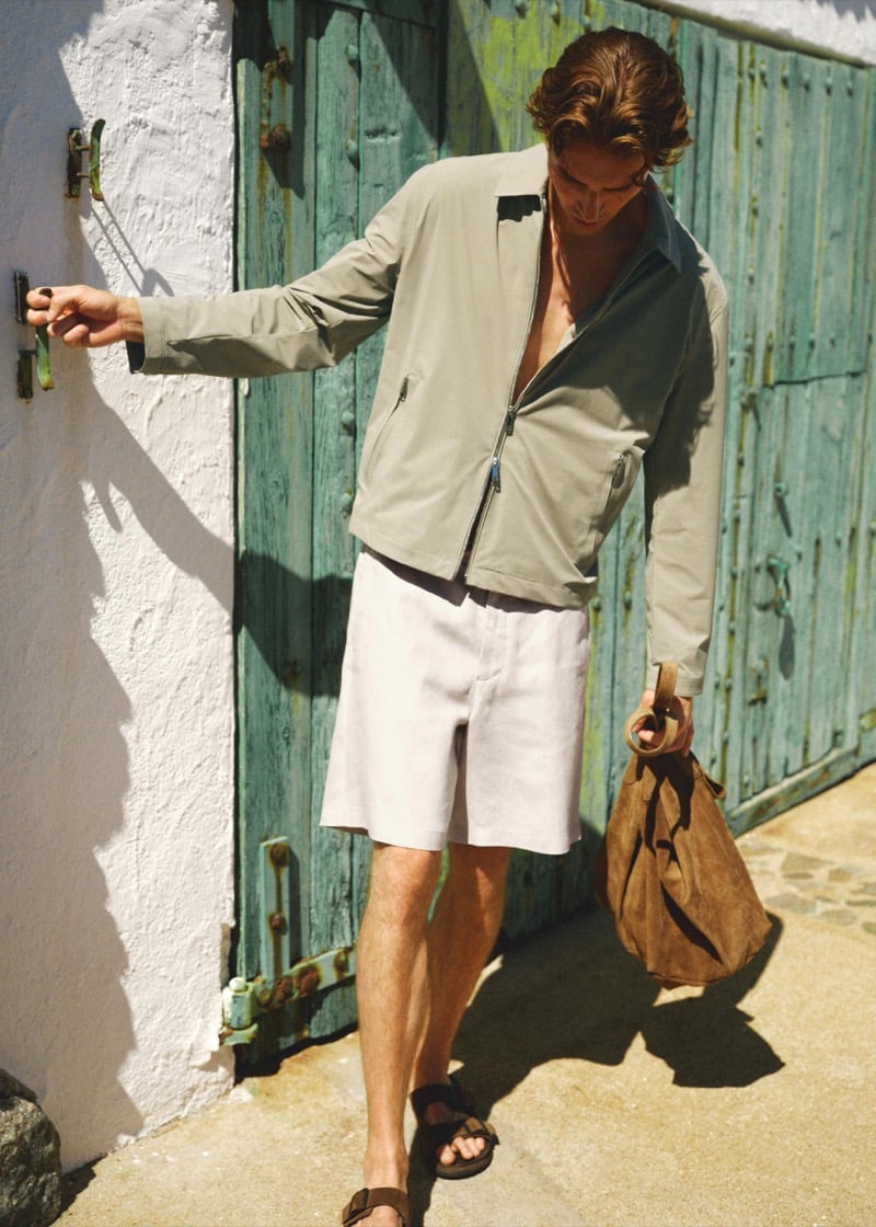 Callum Simpson opens a weathered teal door in an unzipped sage jacket, ivory linen shorts, and a suede tote bag in hand