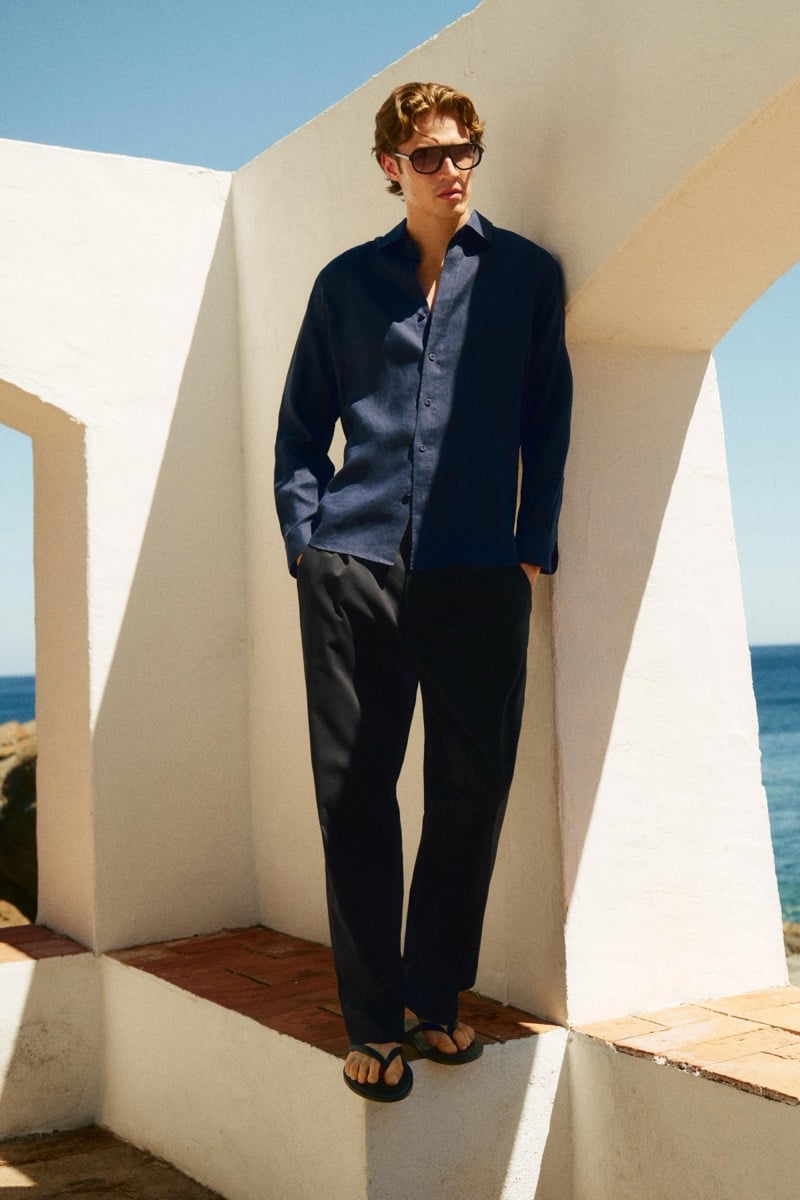 Callum Simpson leans against a whitewashed archway in an open-collar navy shirt, tailored trousers, flip-flop sandals, and sunglasses
