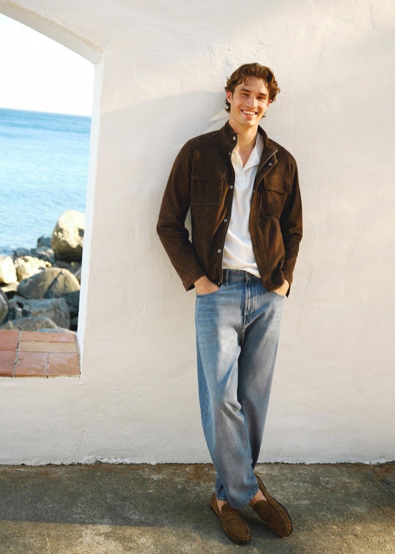 Callum Simpson leans against a whitewashed wall in a chocolate suede jacket over a white polo, light-wash jeans, and suede loafers, smiling toward the sea