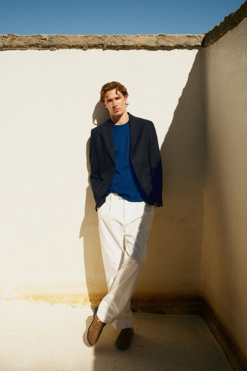 Callum Simpson leans against a sunlit wall in a navy blazer over a cobalt T-shirt, ivory trousers, and suede loafers