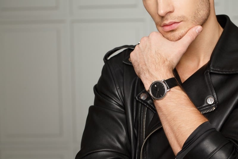 A close-up of a man in a black leather jacket touching his chin while wearing a black watch on a leather strap