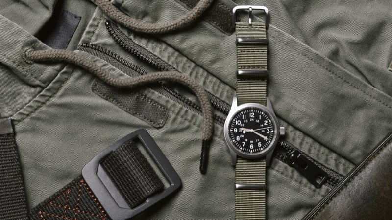 A nylon pass-through strap watch with an olive canvas band rests on a green field jacket