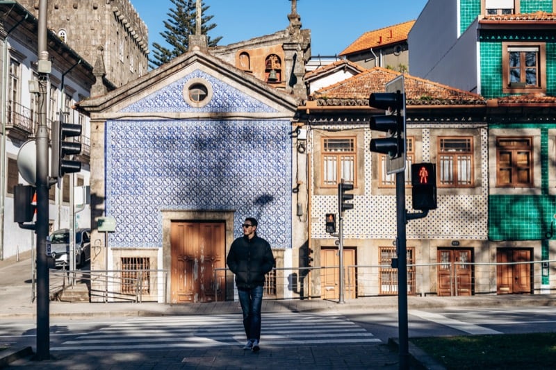 Man in a black puffer jacket walks past a blue tiled chapel facade on a Porto street corner