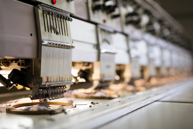 A row of industrial embroidery machines stitching fabric inside a garment factory