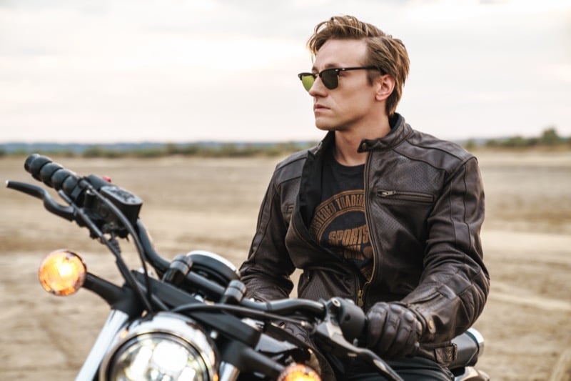 A man on a motorcycle wears a graphic t-shirt under a leather jacket in a desert landscape