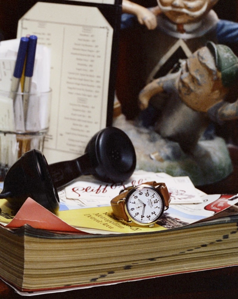 The Shinola Runwell watch with a leather strap rests atop an old phone book beside a vintage rotary telephone receiver and a restaurant menu card for Shinola's "Set the Pace" campaign