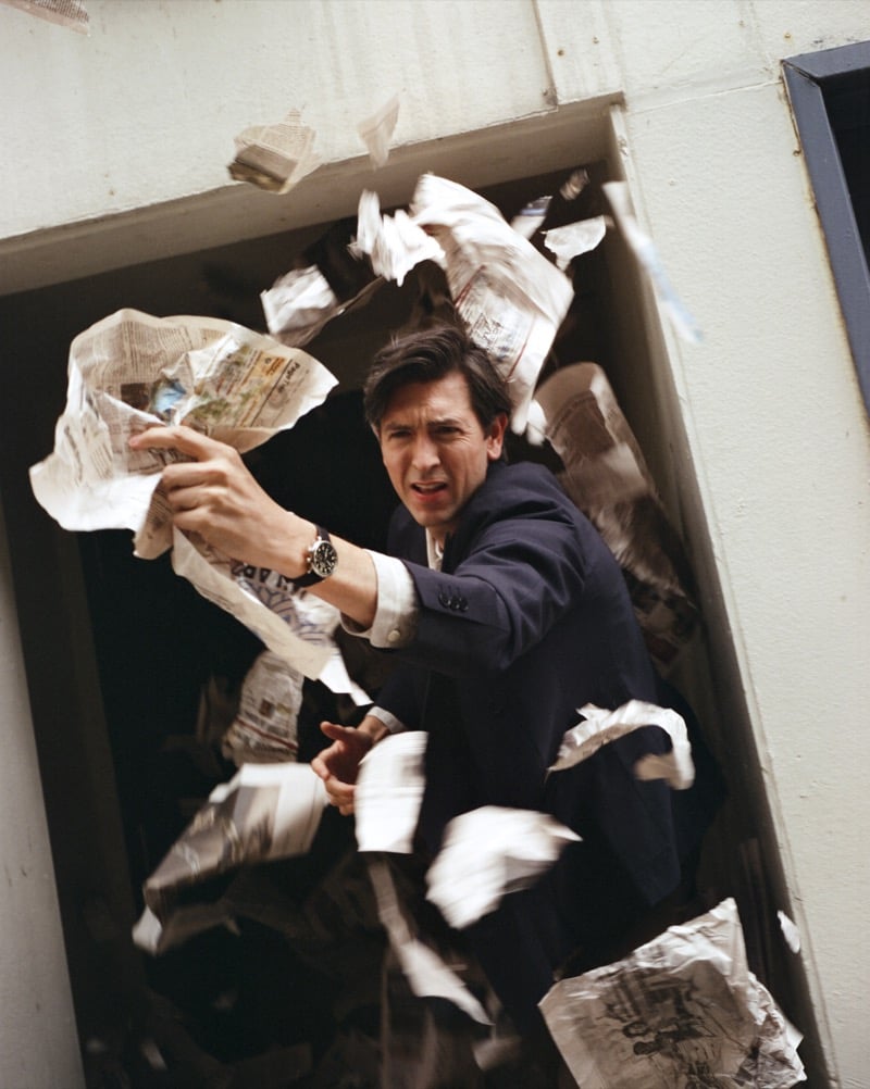 Nicholas Braun bats away a shower of crumpled newspaper pages in a tailored blazer, wearing the Shinola Runwell, in a doorway for Shinola's "Set the Pace" campaign