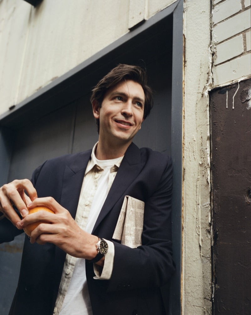 Nicholas Braun smiles in a doorway wearing a tailored blazer over a patterned button-up shirt, holding an orange with a newspaper tucked in his breast pocket, the Shinola Runwell on his wrist, for Shinola's "Set the Pace" campaign
