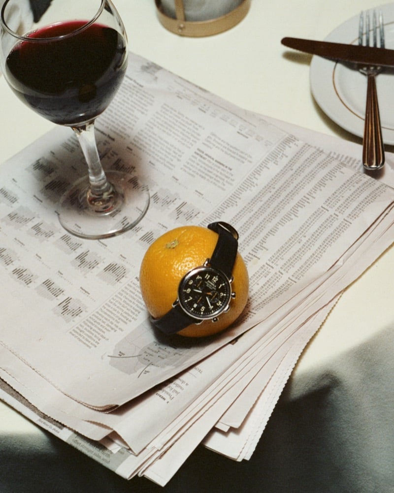 The Shinola Runwell Chronograph rests on an orange set atop a folded newspaper beside a glass of red wine for Shinola's "Set the Pace" campaign