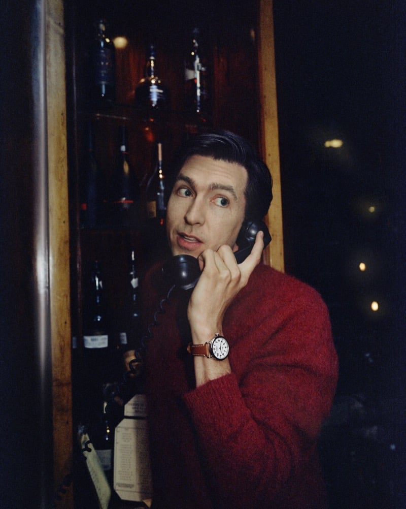 Nicholas Braun in a red knit sweater takes a call on a vintage rotary telephone inside a bottle-lined alcove, the Shinola Runwell on his wrist, for Shinola's "Set the Pace" campaign