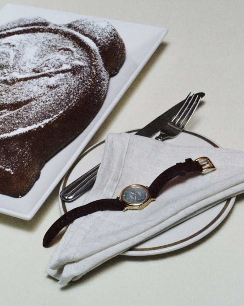 A Shinola Runwell with a blue dial and gold case rests on a linen napkin beside silverware and a powdered-sugar-dusted pastry for Shinola's "Set the Pace" campaign
