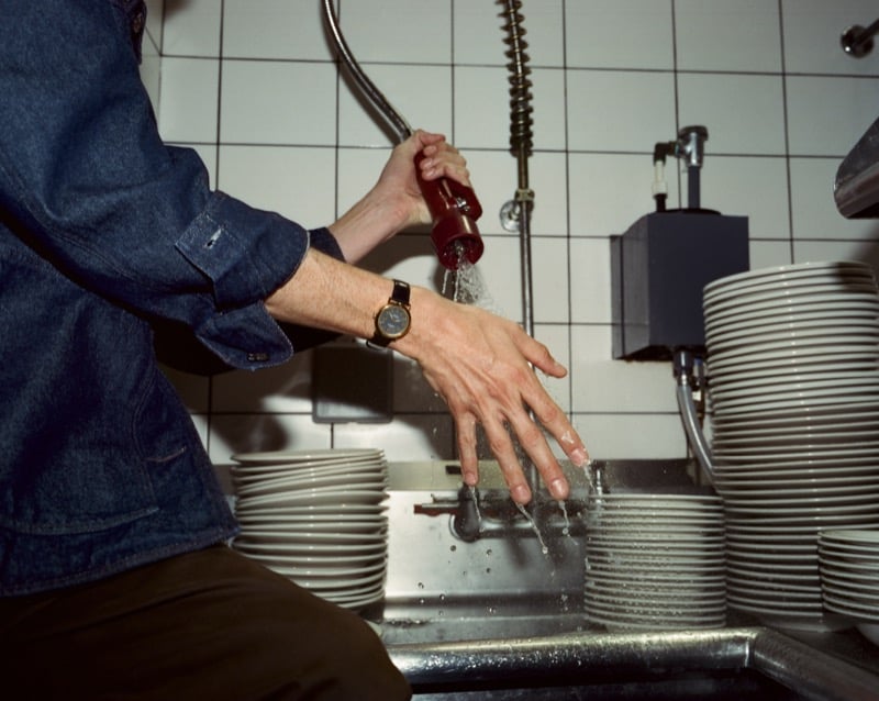 A close-up of Nicholas Braun's hand washing dishes at a restaurant sink, denim shirt sleeve rolled up and the Shinola Runwell Chronograph on his wrist, for Shinola's "Set the Pace" campaign