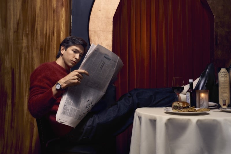 Nicholas Braun reclines in a red knit sweater reading a newspaper with his feet up on a restaurant table set with wine and a burger, wearing the Shinola Runwell, for Shinola's "Set the Pace" campaign