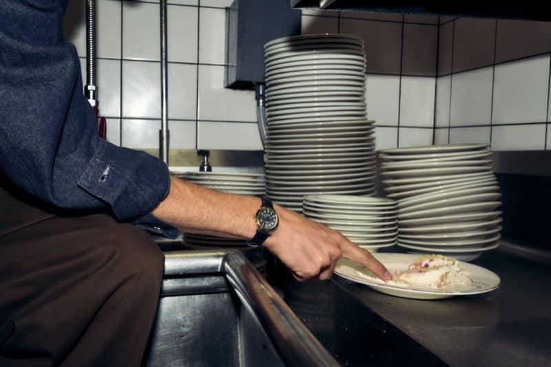 A close-up of Nicholas Braun's hand reaching for a dessert plate in a restaurant kitchen, denim shirt sleeve rolled up and the Shinola Runwell Chronograph on his wrist, for Shinola's "Set the Pace" campaign