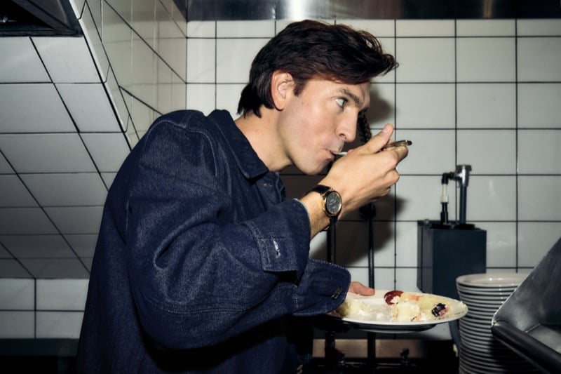 Nicholas Braun in a denim shirt jacket tastes a dish straight from the pan in a restaurant kitchen, the Shinola Runwell Chronograph on his wrist, for Shinola's "Set the Pace" campaign