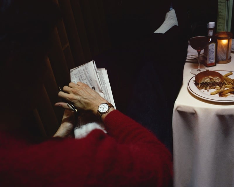 A close-up of Nicholas Braun's hand writing on a folded newspaper in a red sweater sleeve, the Shinola Runwell on his wrist, with a burger and glass of wine on the table behind him, for Shinola's "Set the Pace" campaign