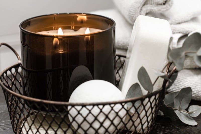Lit candle, bar soap, and rolled towels arranged in a wire basket with a eucalyptus sprig