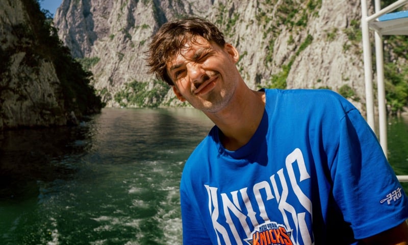A man smiles on a boat wearing a blue team t-shirt with mountains in the background