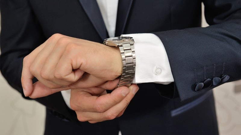 A man in a tuxedo adjusts a stainless steel bracelet watch at his wrist