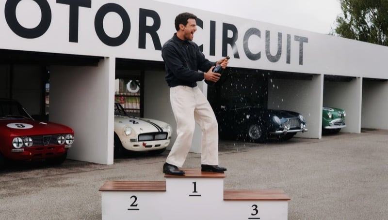 Theo James pops champagne on the winner's podium at Goodwood Motor Circuit, wearing a dark knit, light trousers, and Church's dress shoes, with vintage racers lined up behind him