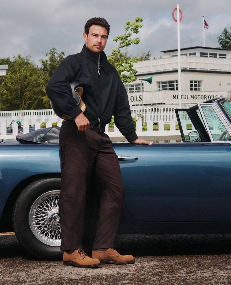 Theo James leans against a vintage Aston Martin at Goodwood, a helmet tucked under one arm, wearing a black jacket, brown trousers, and tan suede Church's shoes