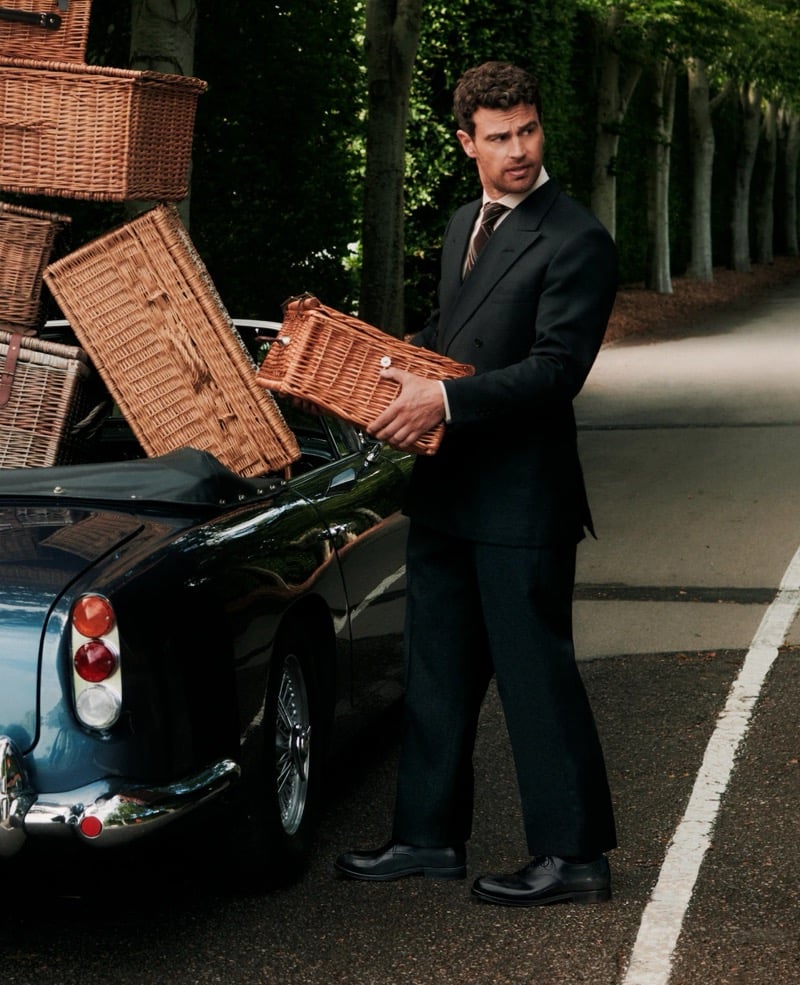 Theo James in a dark double-breasted suit and tie unloads wicker picnic baskets from a vintage Aston Martin on a tree-lined country road
