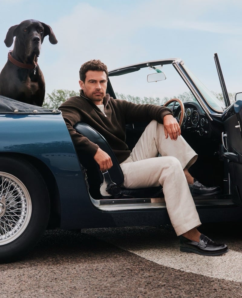 Theo James sits behind the wheel of a vintage Aston Martin convertible with a Great Dane perched in the back seat, wearing a brown knit sweater and black Church's loafers