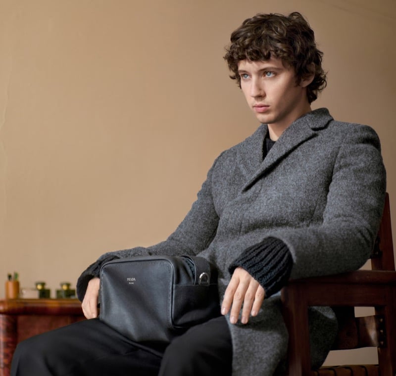 Troye Sivan sits in a wood chair wearing a gray wool coat with a knit cuff, holding a brown leather Prada bag in his lap