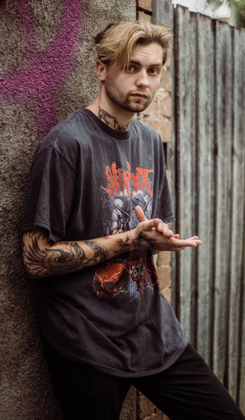 A man with tattooed arms wears a faded black band t-shirt against a graffiti wall