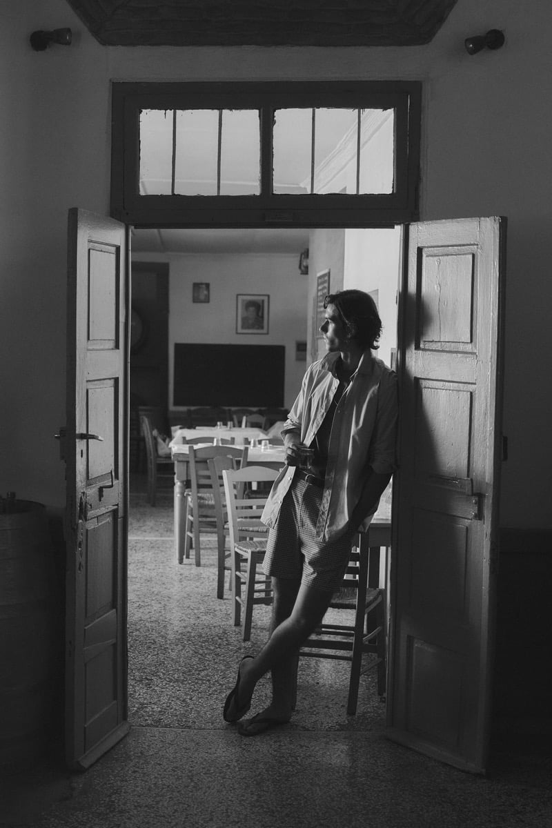 Black-and-white image of Tony Özkan framed in a taverna doorway, wearing an open shirt, checked shorts, and sandals