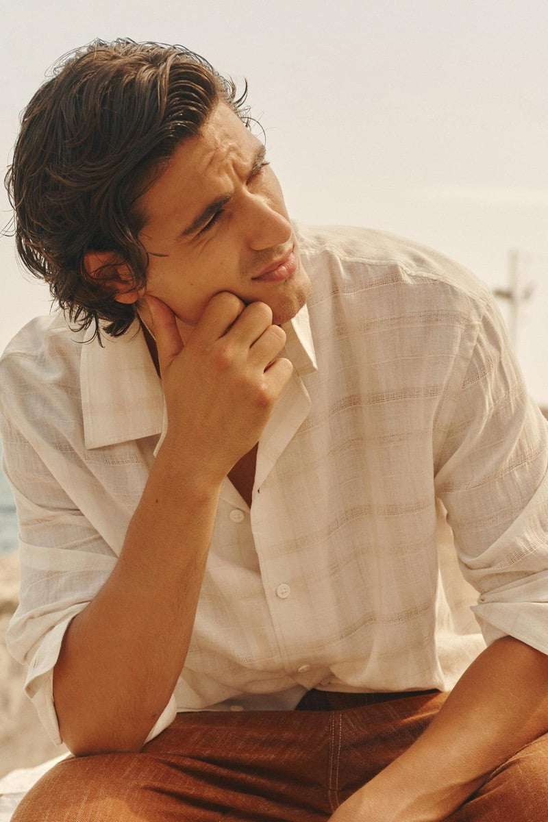 Tony Özkan rests his chin on his hand in an unbuttoned woven shirt and terracotta trousers by a rocky Athens shoreline