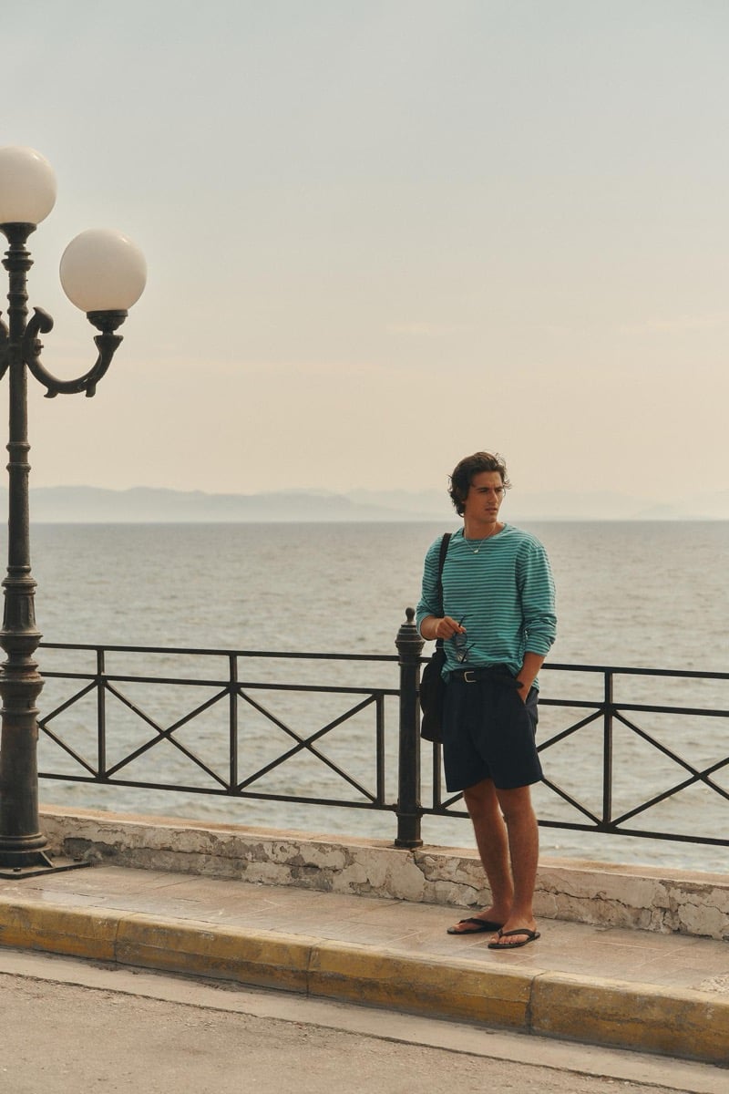 Tony Özkan stands along a waterfront railing in a green and blue striped top and dark shorts, with the sea behind him
