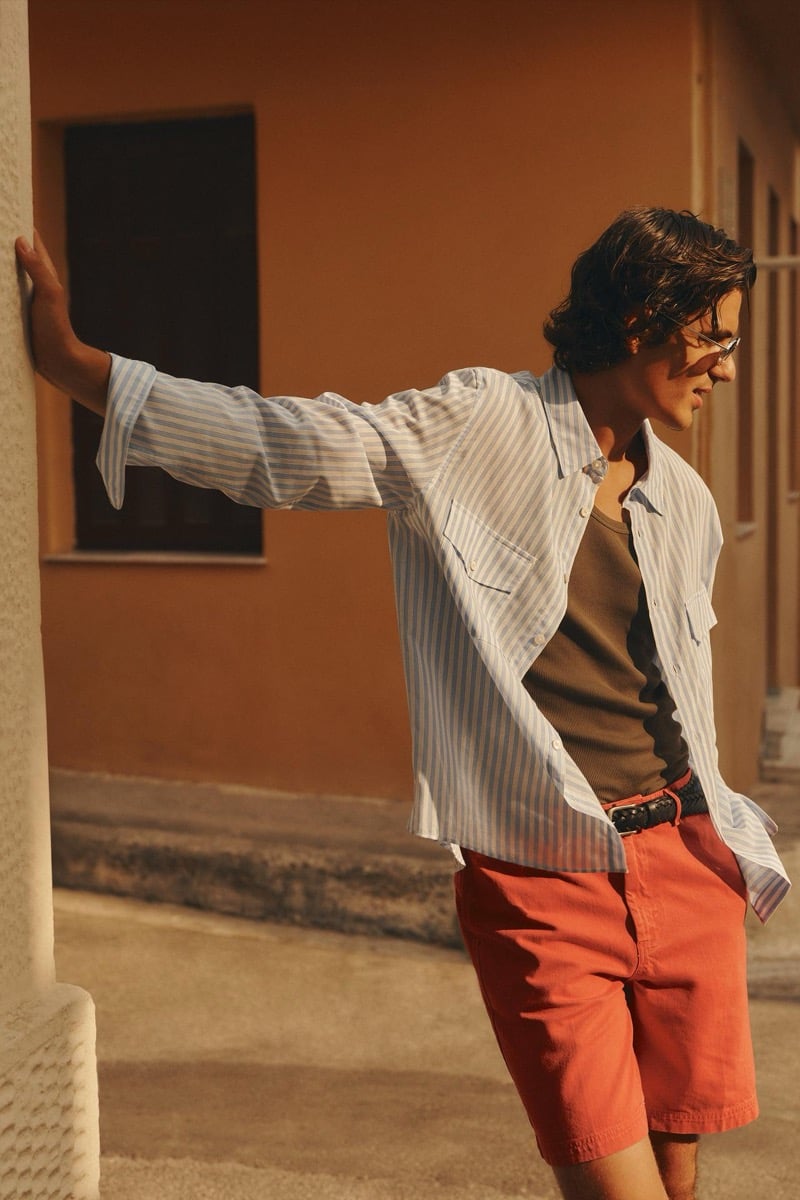 Tony Özkan stands against a terracotta wall in a pale blue striped overshirt, an olive tank top, and coral shorts