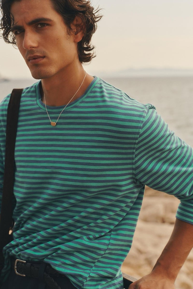 Close-up of Tony Özkan in a green and blue striped knit top with a pendant necklace against the Aegean Sea