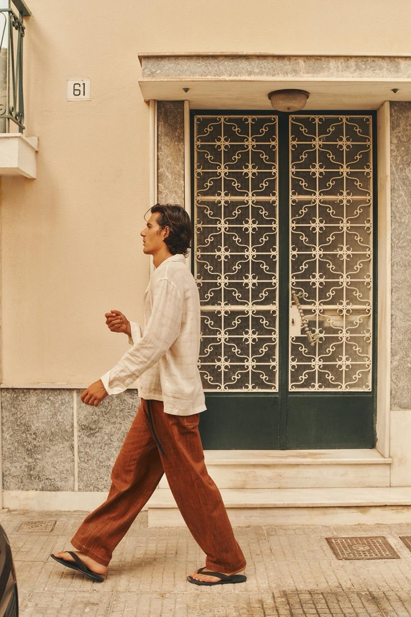 Tony Özkan walks past an ornate green door in a woven shirt and terracotta trousers, sandals on his feet