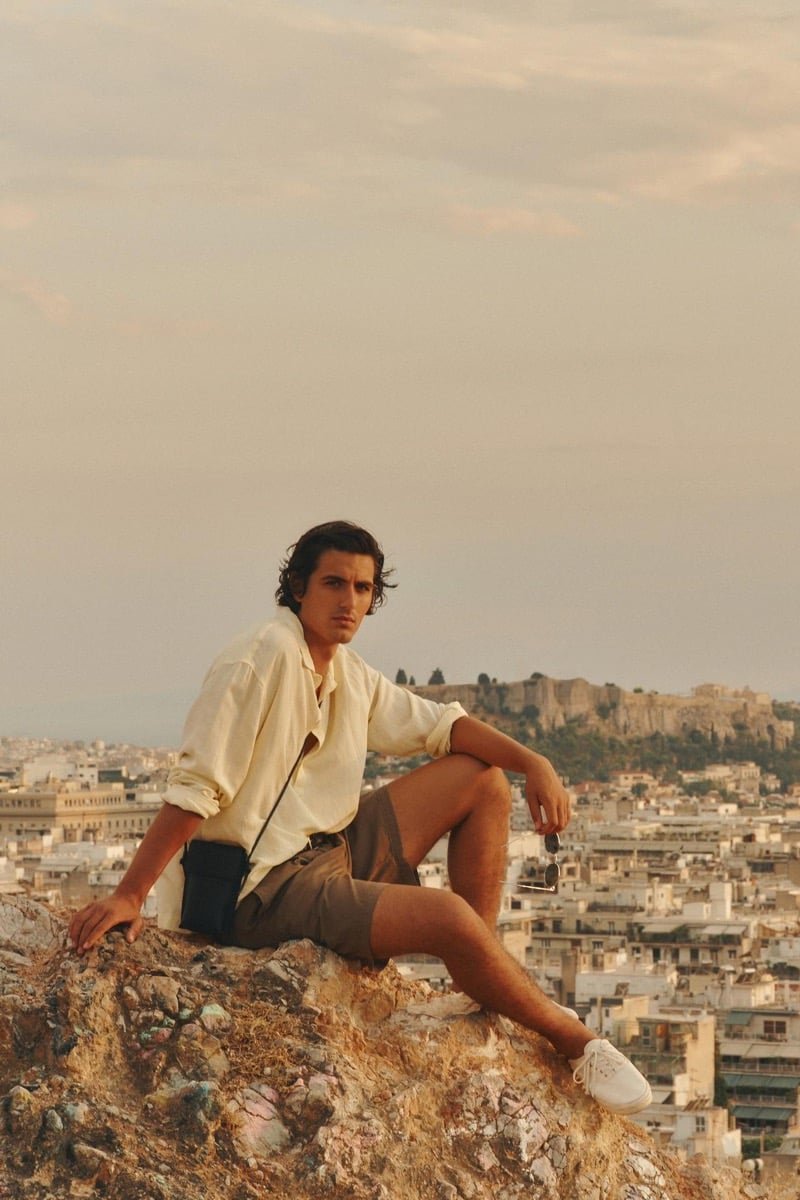 Tony Özkan sits on a rocky hillside in a pale shirt and tan shorts with the Acropolis rising behind him