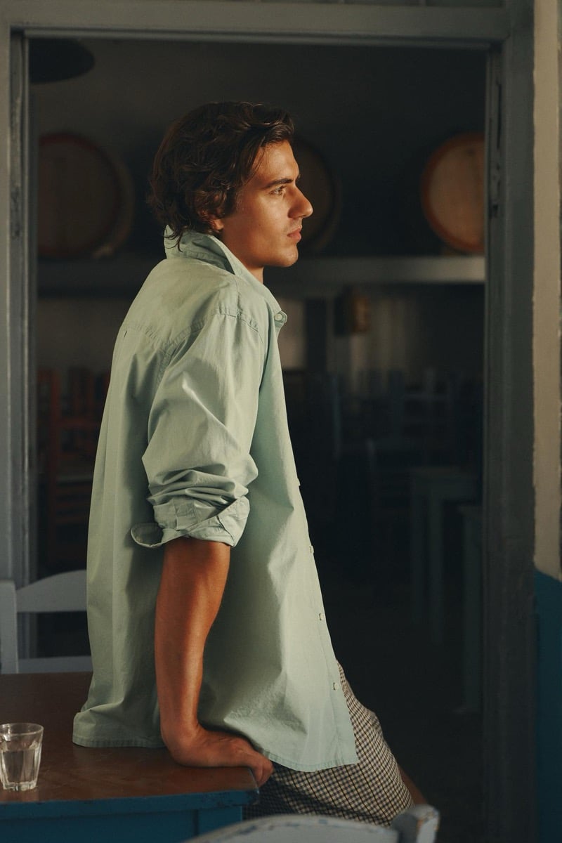 Tony Özkan sits at a table in a sage green shirt, a taverna interior with wine barrels visible behind him