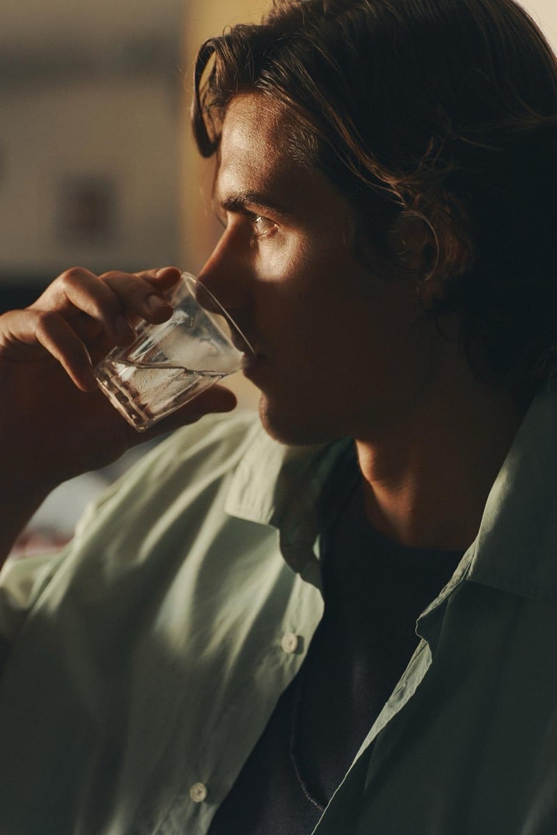 Close-up of Tony Özkan drinking from a glass in a sage green shirt, sunlight catching his face