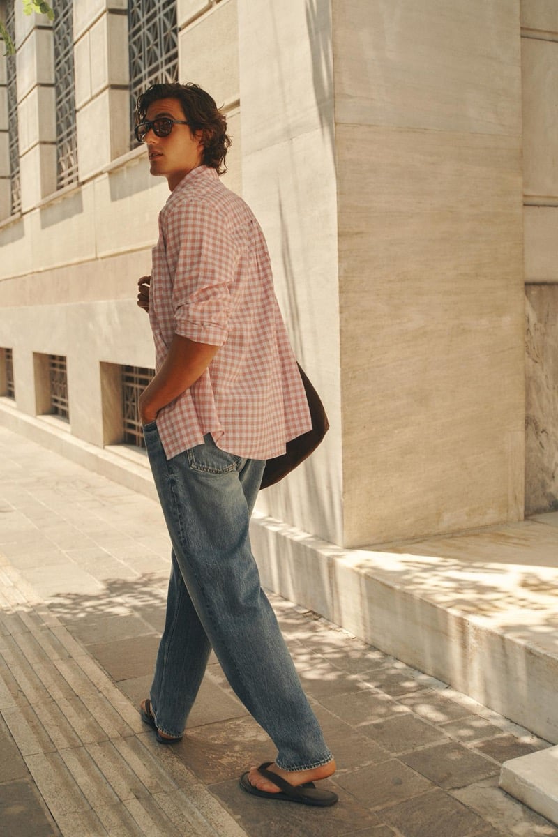 Tony Özkan walks along a stone building in a pink checked shirt, loose denim jeans, and sandals, a dark bag over his shoulder