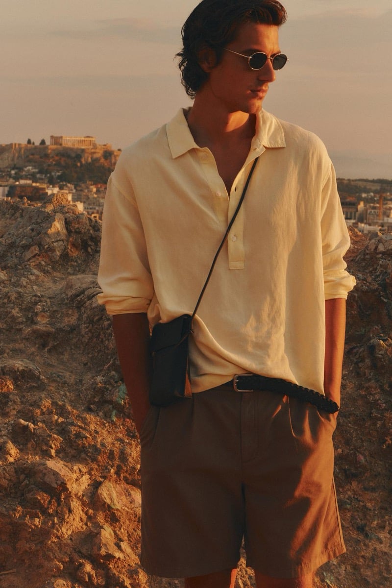 Tony Özkan stands on a hillside in a pale yellow popover shirt and tan shorts with the Acropolis visible in the distance