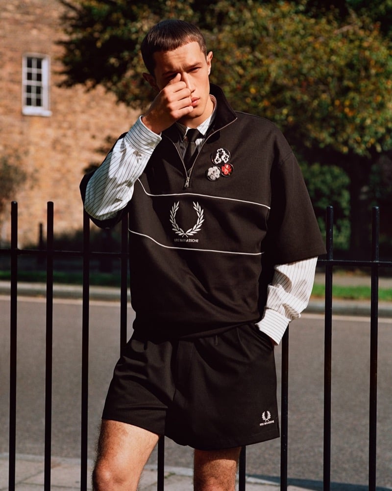 Achille Genet leans on iron railings in a black pique half-sleeve zip top with Fred Perry laurel embroidery and enamel pins, layered over a pinstripe shirt with shorts