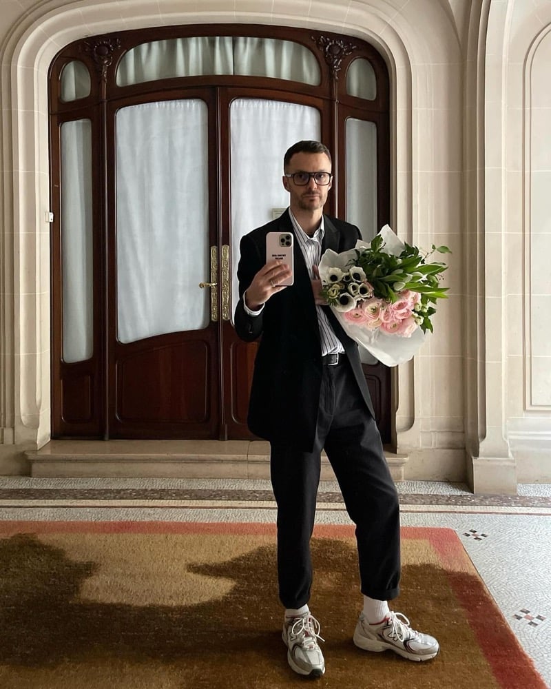 Kris Van Assche takes a mirror selfie in a black blazer and cropped trousers, holding a large bouquet, in an art nouveau interior