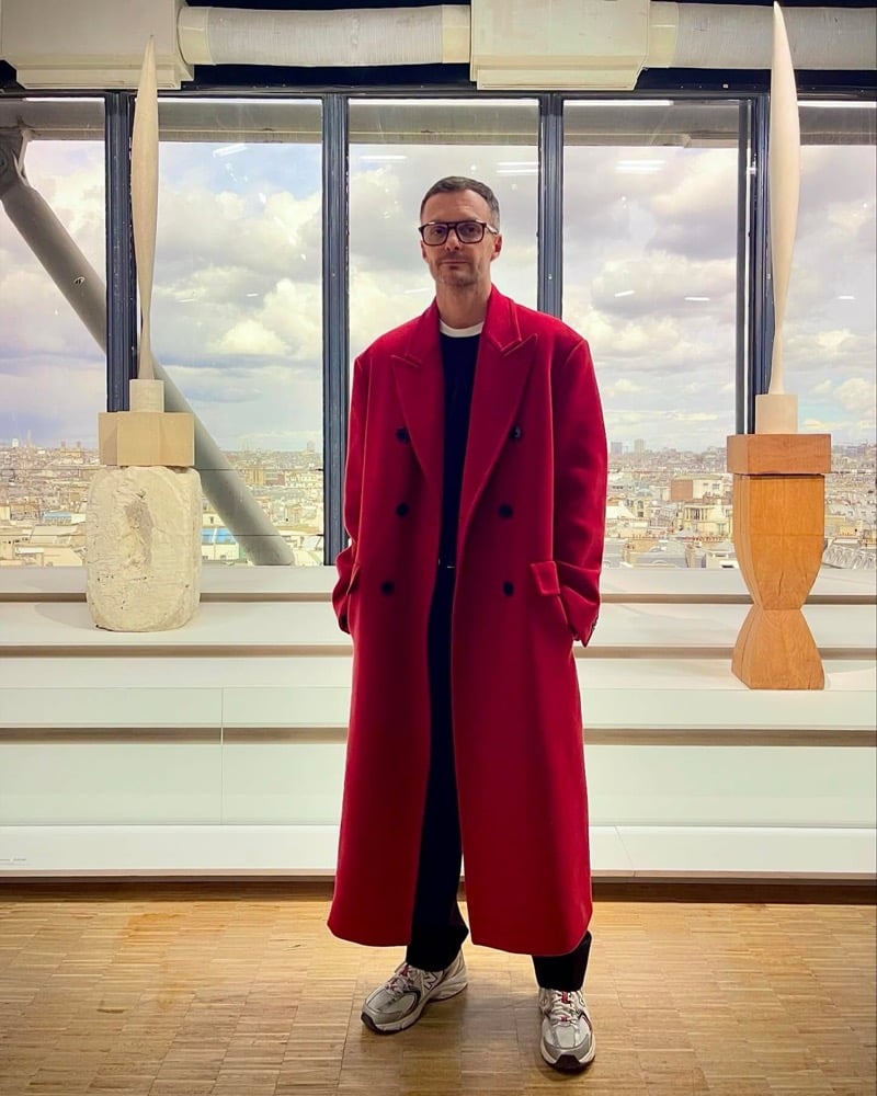 Kris Van Assche stands in a long red double-breasted overcoat over dark clothes and sneakers in a studio with a Paris rooftop view
